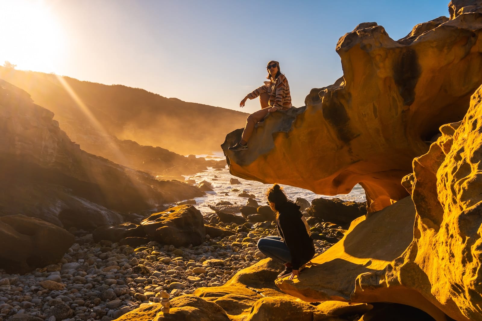 Two people sit and crouch on rugged orange rocks along a sunlit coastline at golden hour, with a dramatic cliff backdrop and sea below.