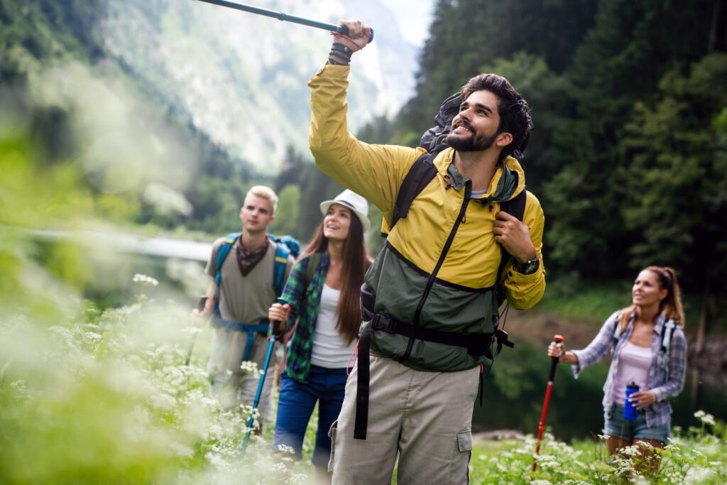 Four hikers pose for a photo in a forested area by a lake; man in a yellow jacket raises a trekking pole as others watch.