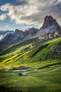 Tall jagged mountain peak rises above a green-alpine valley with a few houses and a winding road in the foreground.